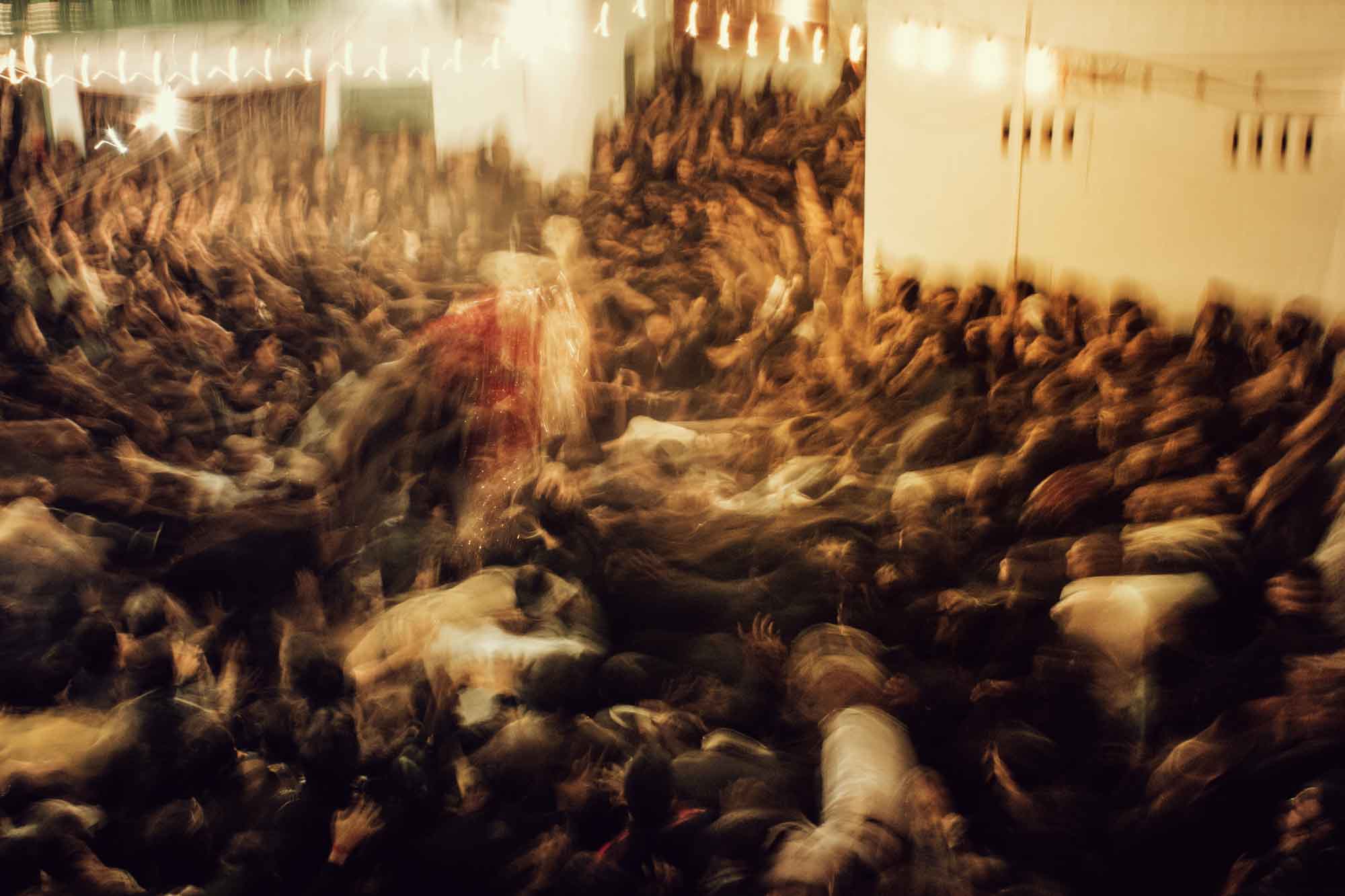 A crowd of mourners blurred in motion beneath strung lights, from Horse / Men by Nad-E-Ali